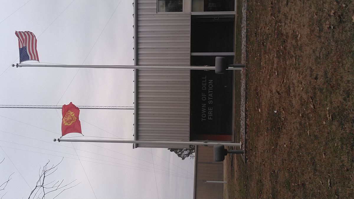 Picture of Dell Fire Dept sign with two flag poles in the front with the US Flag and Fire Dept flag flying.