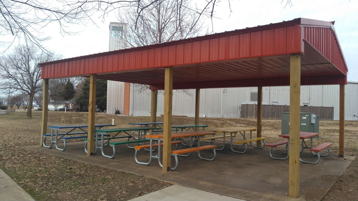 A red roofed pavillion in Dell Park with seven picket table and bench combos under it