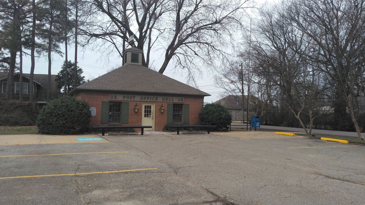 Dell post office building with a flag pole to the right and empty parking lot.